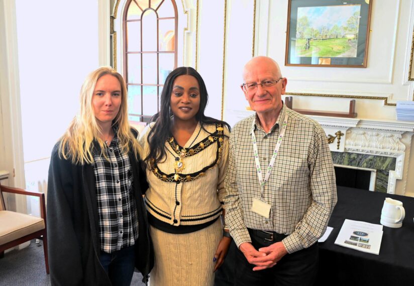Three people stand together in front of a fireplace in a bright room. They are smiling slightly, creating a warm and welcoming atmosphere.