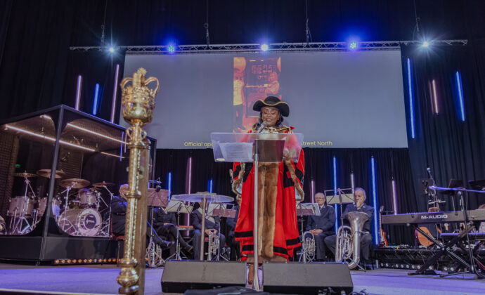 A person in ceremonial attire, including a feathered hat and red robe, speaks at a podium. A band is in the background, and a large screen is above. The setting is formal and grand.