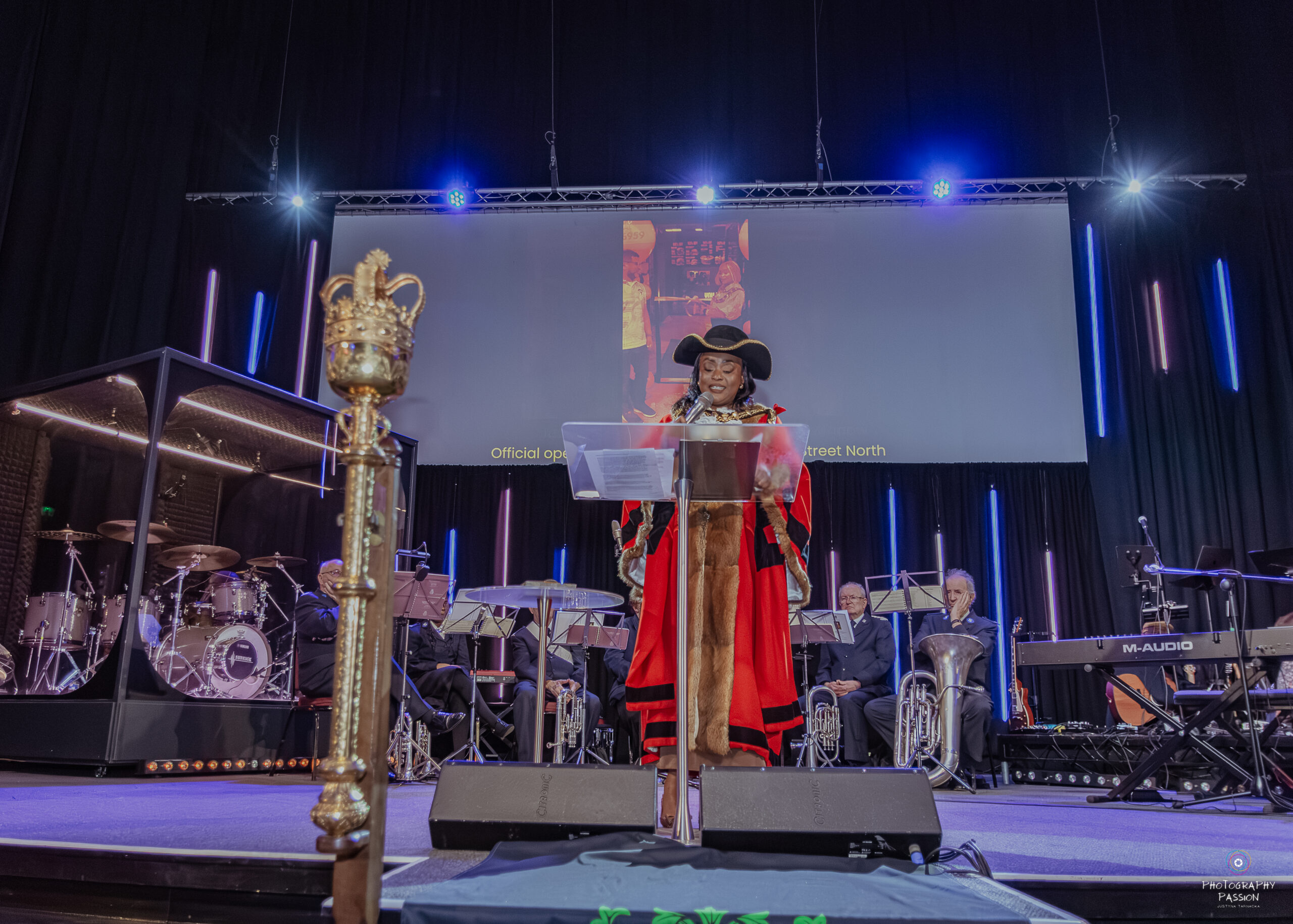 A person in ceremonial attire, including a feathered hat and red robe, speaks at a podium. A band is in the background, and a large screen is above. The setting is formal and grand.