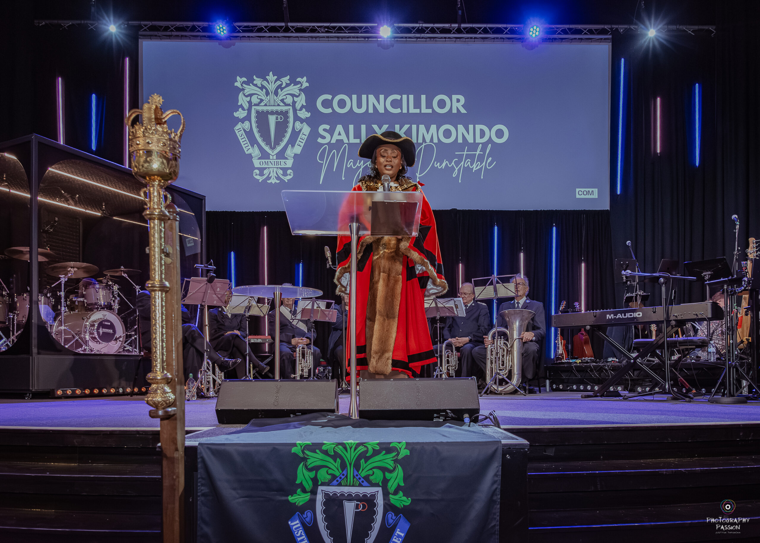 Councillor in ceremonial red robe and hat speaks at a podium on stage. A band with instruments is seated behind, with blue lights highlighting the scene.