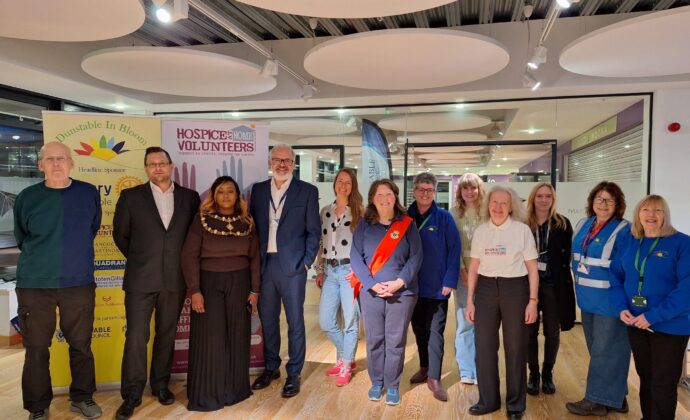 A diverse group of 12 people stands in a well-lit room, smiling. Two banners in the background read 