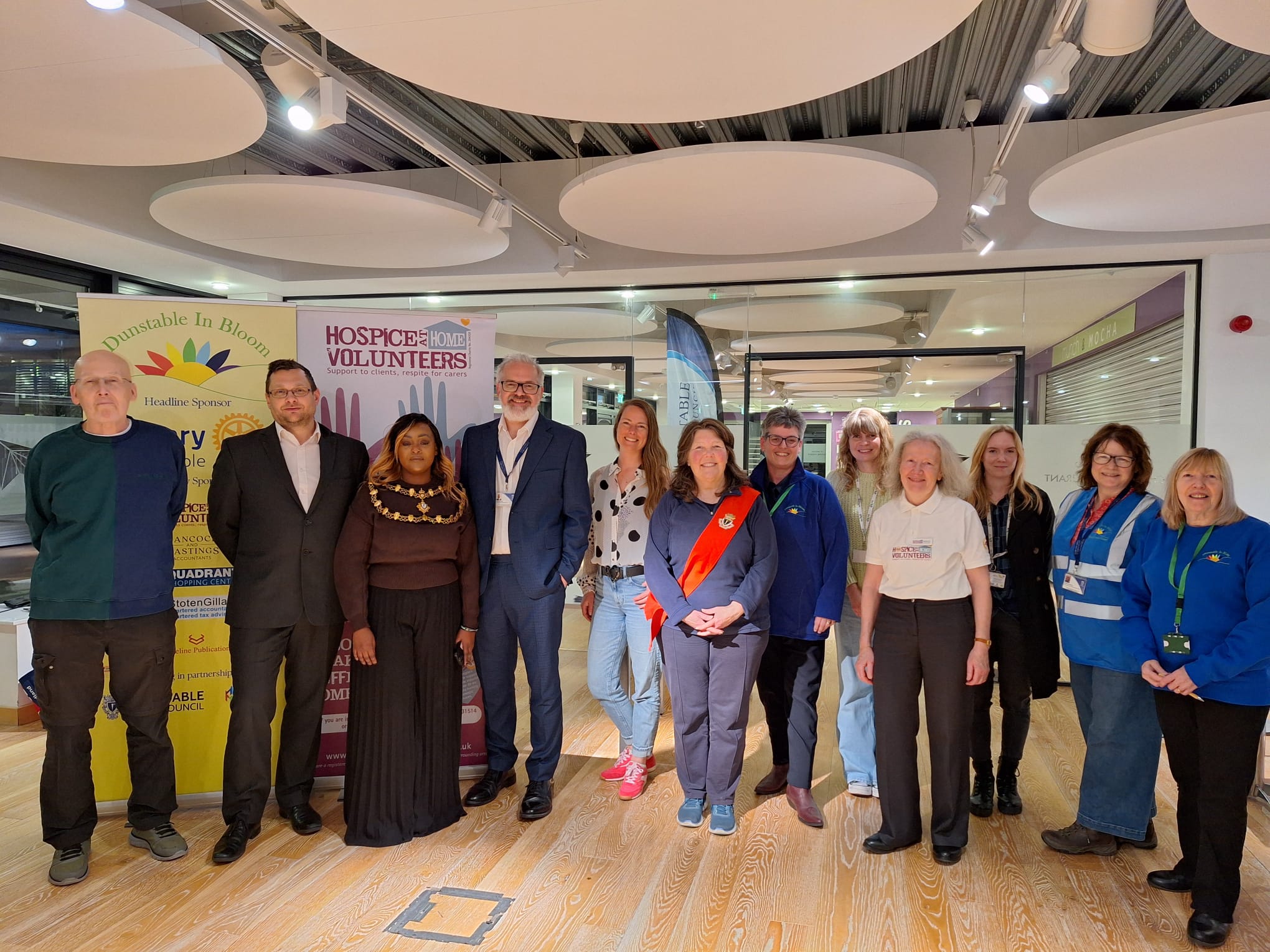 A diverse group of 12 people stands in a well-lit room, smiling. Two banners in the background read 