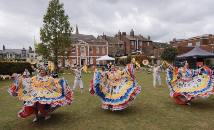 Four dancers in colorful traditional dresses perform a folkloric dance outdoors on a grassy area, with historic buildings and onlookers in the background.