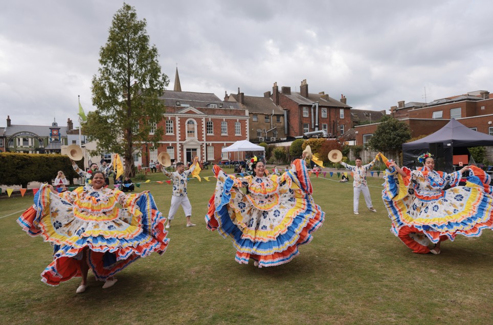 Four dancers in colorful traditional dresses perform a folkloric dance outdoors on a grassy area, with historic buildings and onlookers in the background.