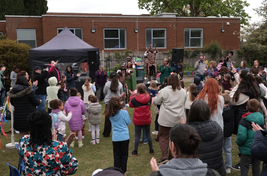 A crowd of people, including children and adults, gather outdoors in front of a stage with musicians performing, next to a brick building.