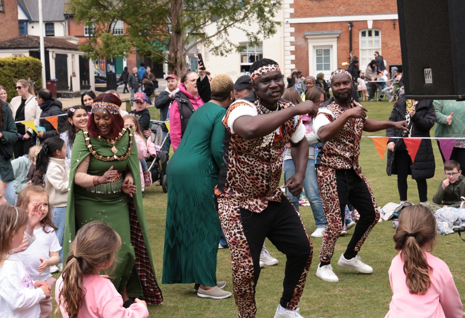 A lively outdoor event with two dancers in leopard print outfits performing energetically. A crowd watches, smiling, with children joining in.