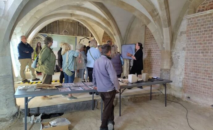 A group of people gathered in a vaulted stone room, listening to a woman presenting information next to a monitor.
