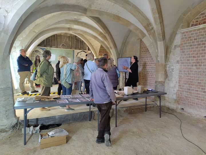 A group of people gathered in a vaulted stone room, listening to a woman presenting information next to a monitor.