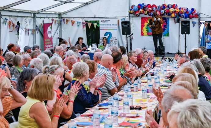 A large group of people sit at long tables under a tent, clapping while a man singer on stage