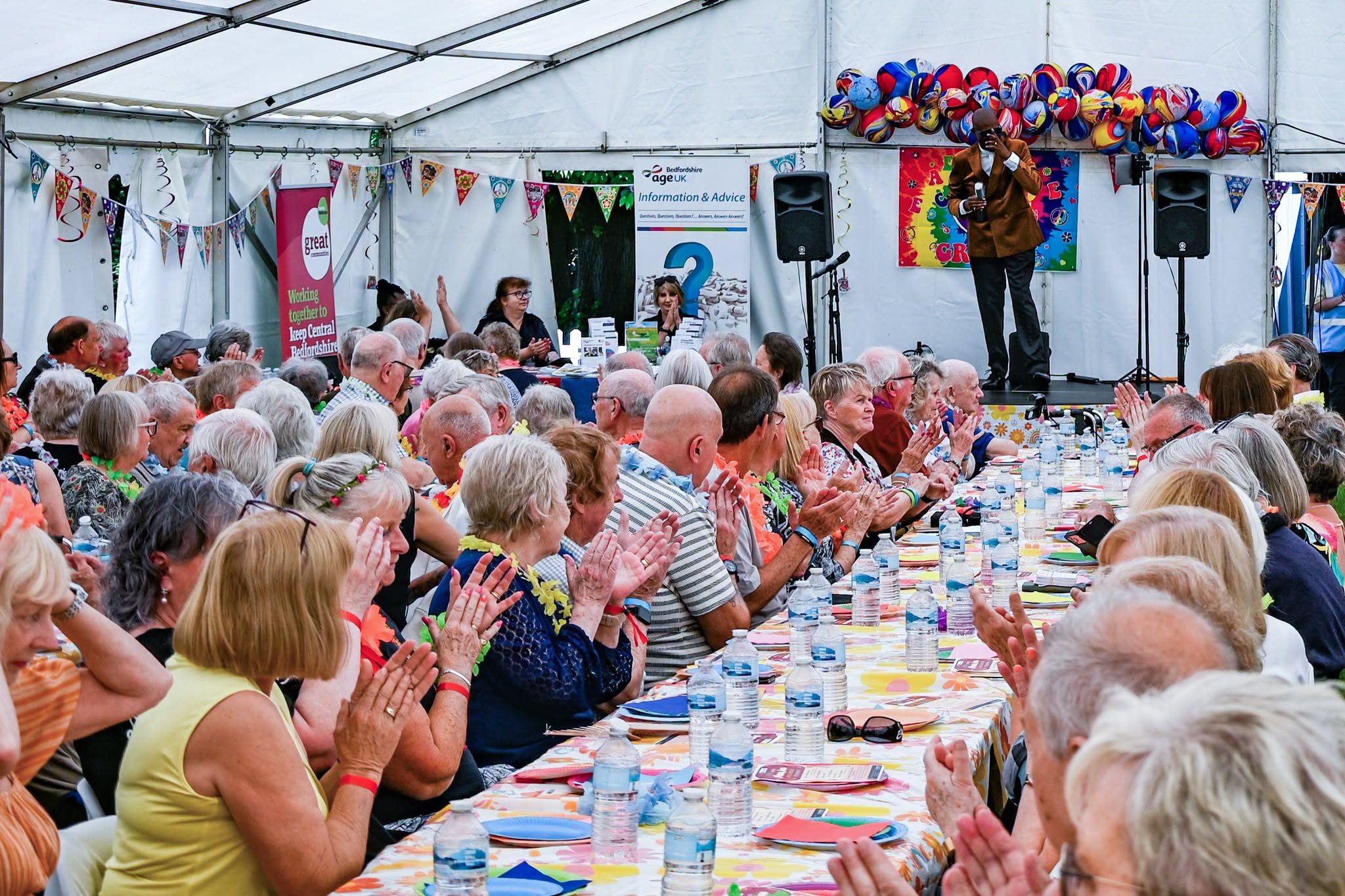A large group of people sit at long tables under a tent, clapping while a man singer on stage