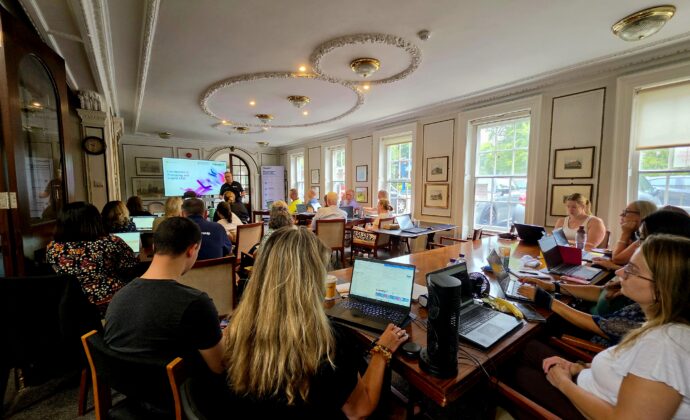 Lively workshop in a sunlit room, with participants using laptops around tables while a presenter speaks beside a projected screen.