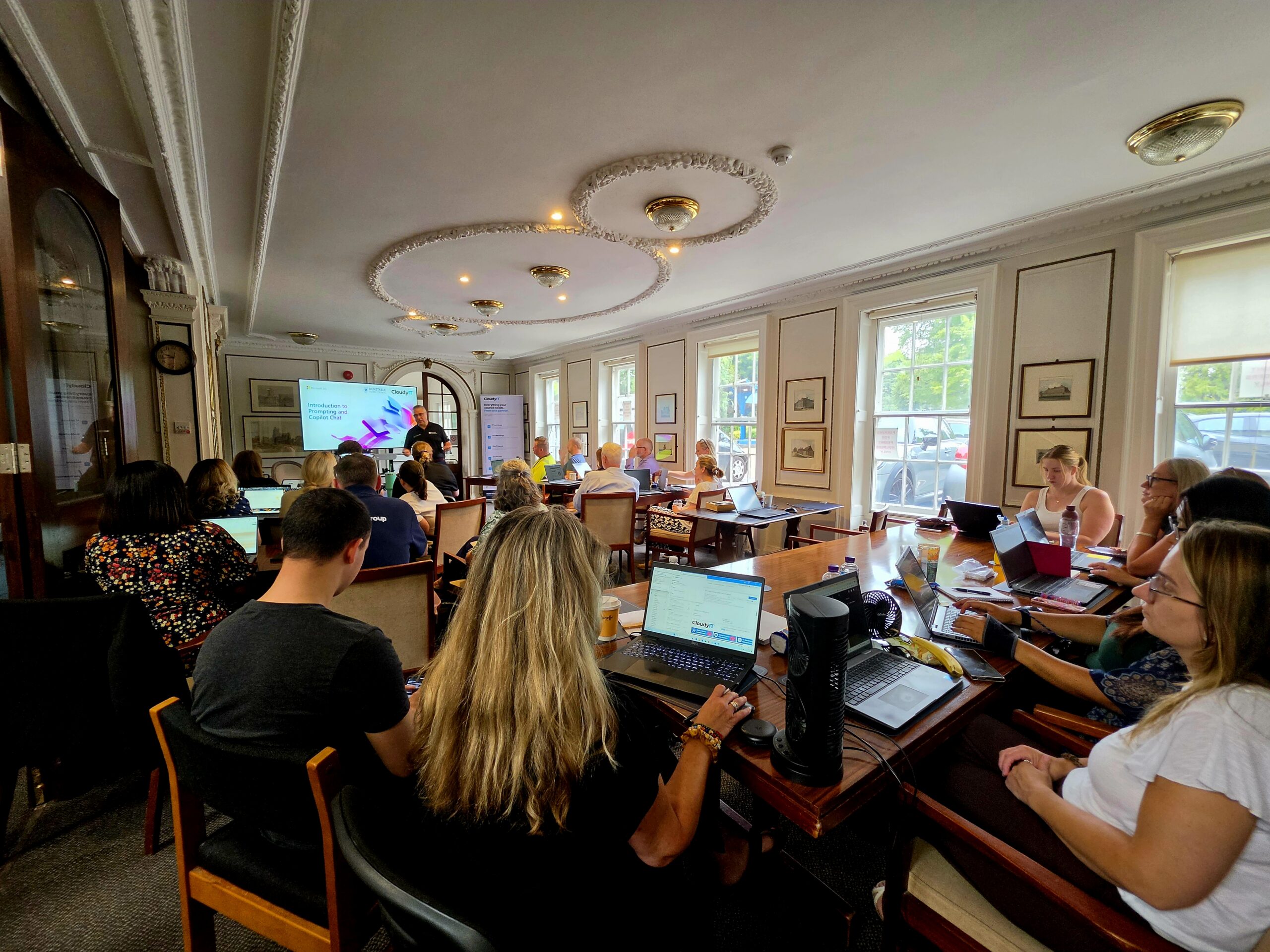 Lively workshop in a sunlit room, with participants using laptops around tables while a presenter speaks beside a projected screen.