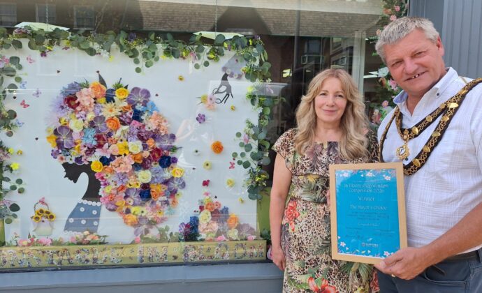 Two people stand in front of a decorated window with a colorful floral art display; one holds a framed certificate or award, both smiling at the camera.