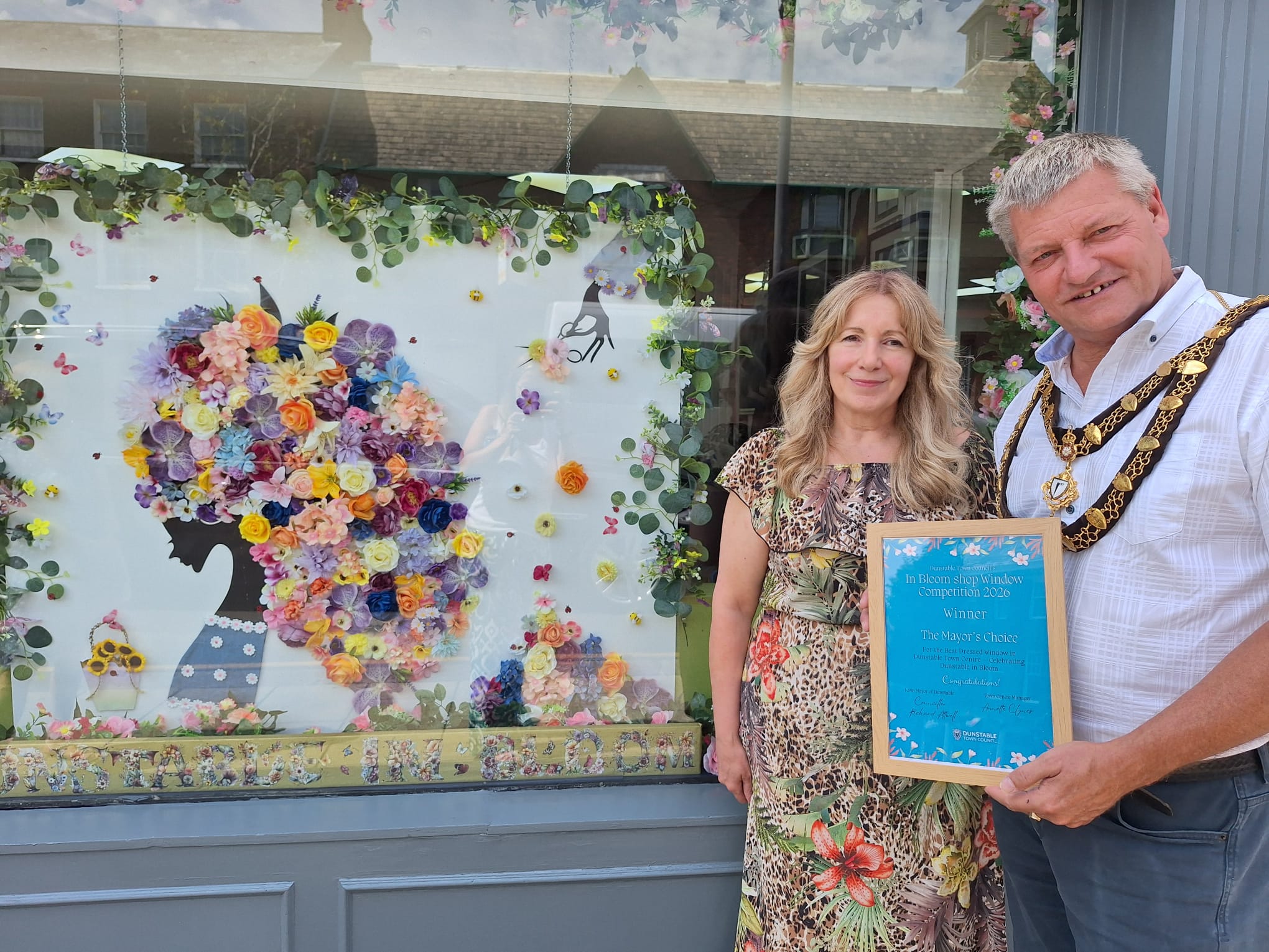 Two people stand in front of a decorated window with a colorful floral art display; one holds a framed certificate or award, both smiling at the camera.
