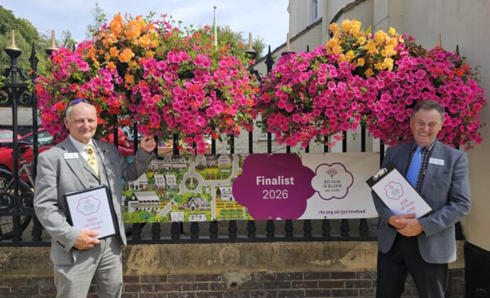 Two men in suits stand beside colorful hanging flower baskets and a sign that reads “Finalist 2026, Britain in Bloom.” Both are holding folders and smiling at the camera.
