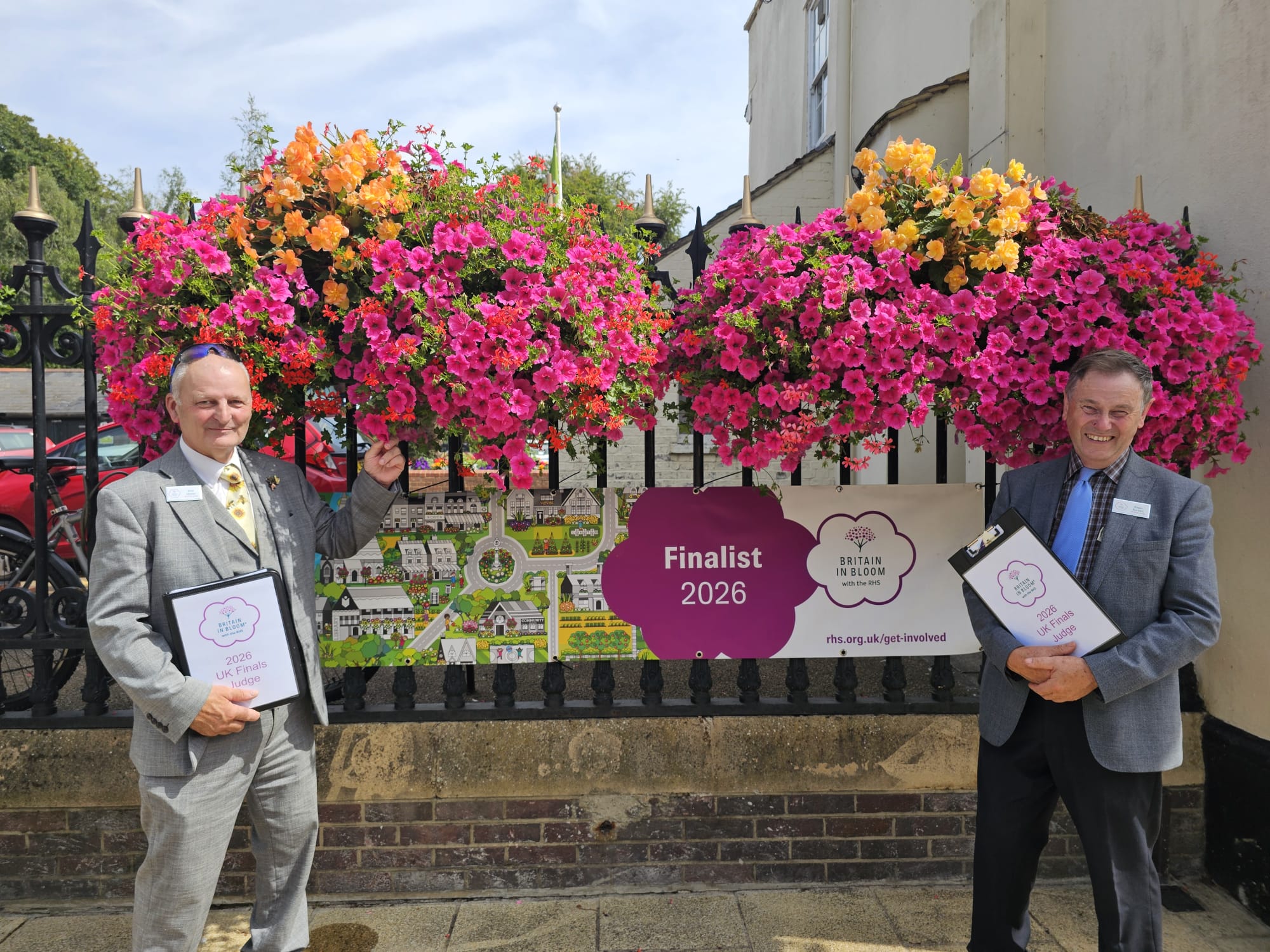 Two men in suits stand beside colorful hanging flower baskets and a sign that reads “Finalist 2026, Britain in Bloom.” Both are holding folders and smiling at the camera.