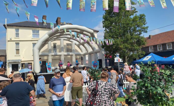 Crowd enjoys a lively outdoor street festival with colorful bunting, a white inflatable stage, music, stalls, and sunny skies.