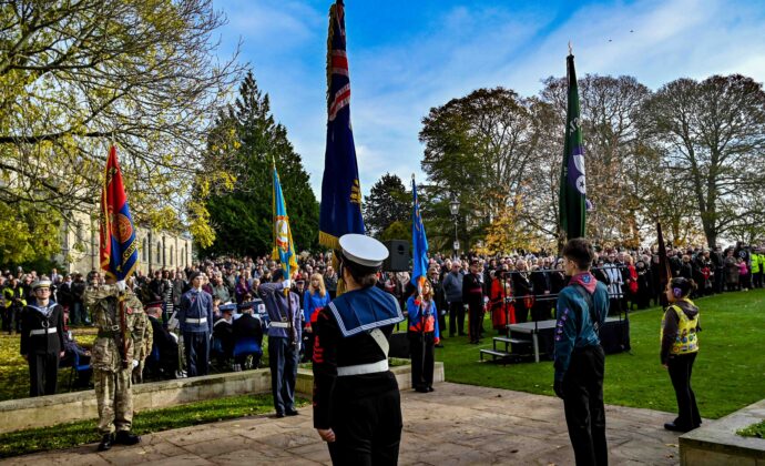 Solemn outdoor remembrance ceremony in a park, with uniformed participants holding colorful flags before a large crowd beneath blue skies.