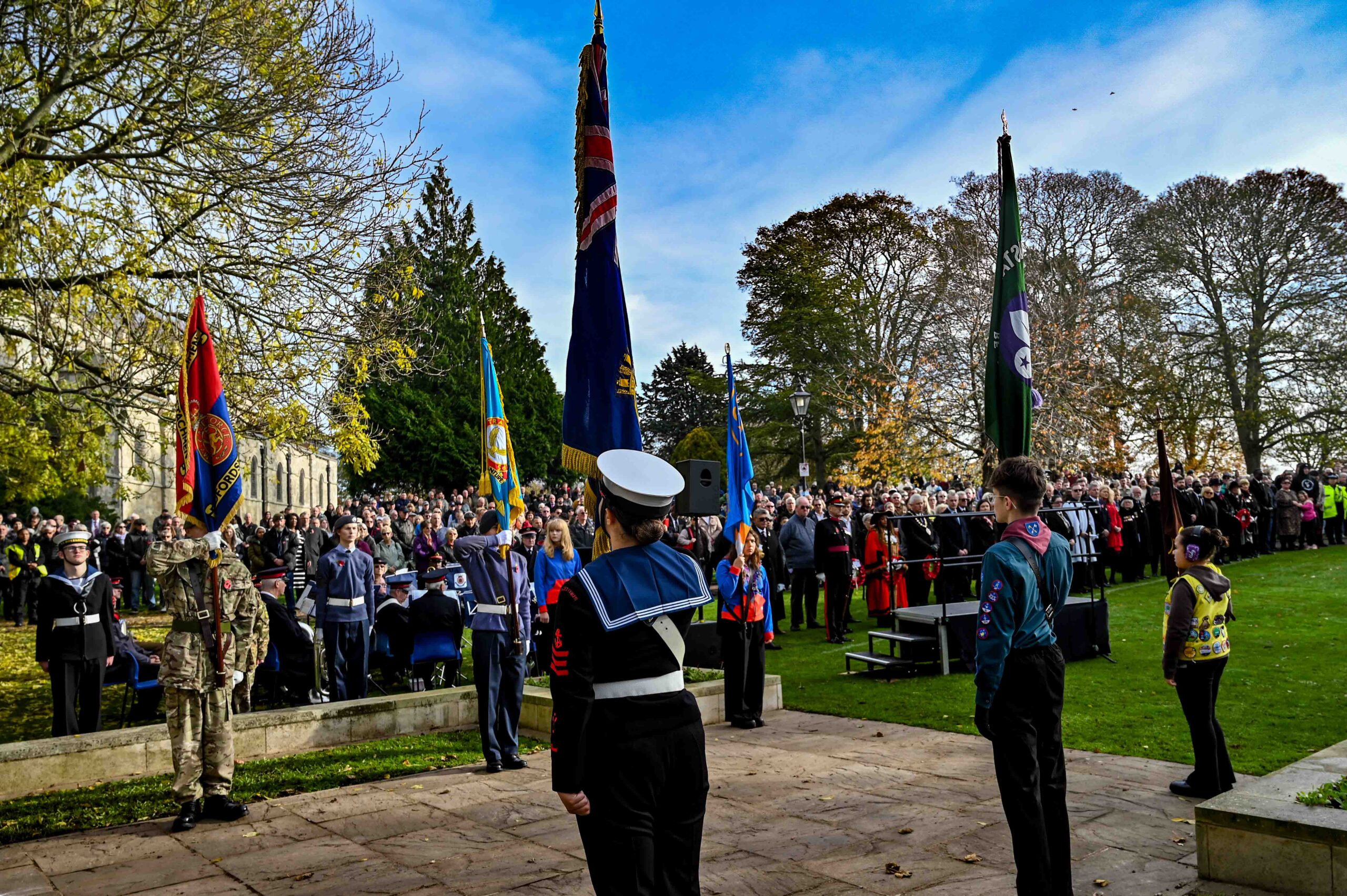 Solemn outdoor remembrance ceremony in a park, with uniformed participants holding colorful flags before a large crowd beneath blue skies.
