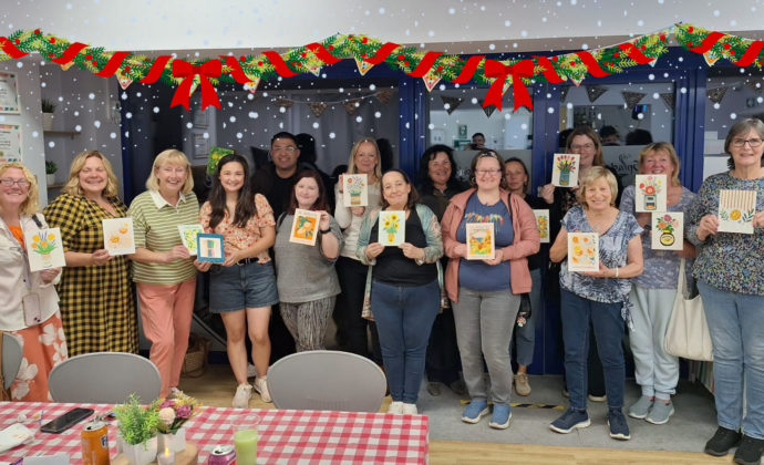 A group of people stands indoors, smiling and holding up handmade cards, with bunting decorations and tables with checked tablecloths in the foreground.