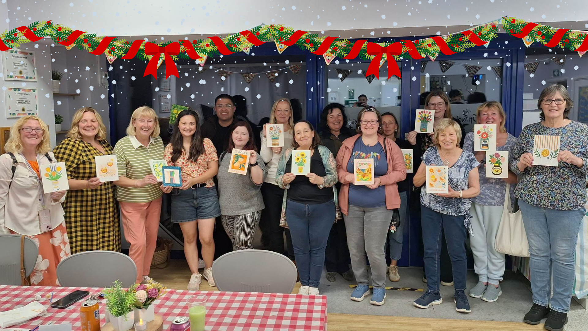 A group of people stands indoors, smiling and holding up handmade cards, with bunting decorations and tables with checked tablecloths in the foreground.