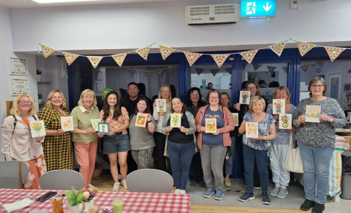 A group of people stands indoors, smiling and holding up handmade cards, with bunting decorations and tables with checked tablecloths in the foreground.
