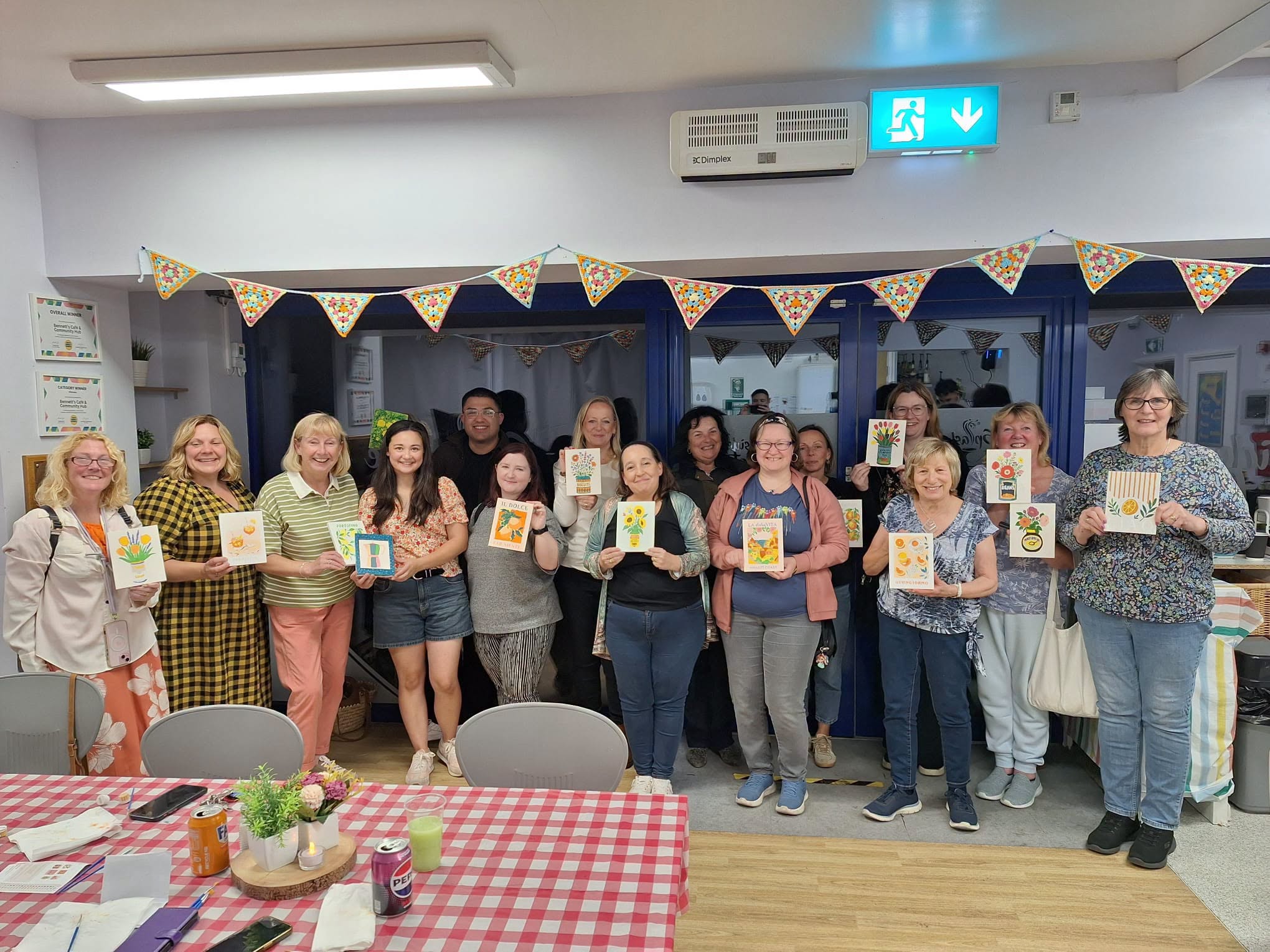 A group of people stands indoors, smiling and holding up handmade cards, with bunting decorations and tables with checked tablecloths in the foreground.